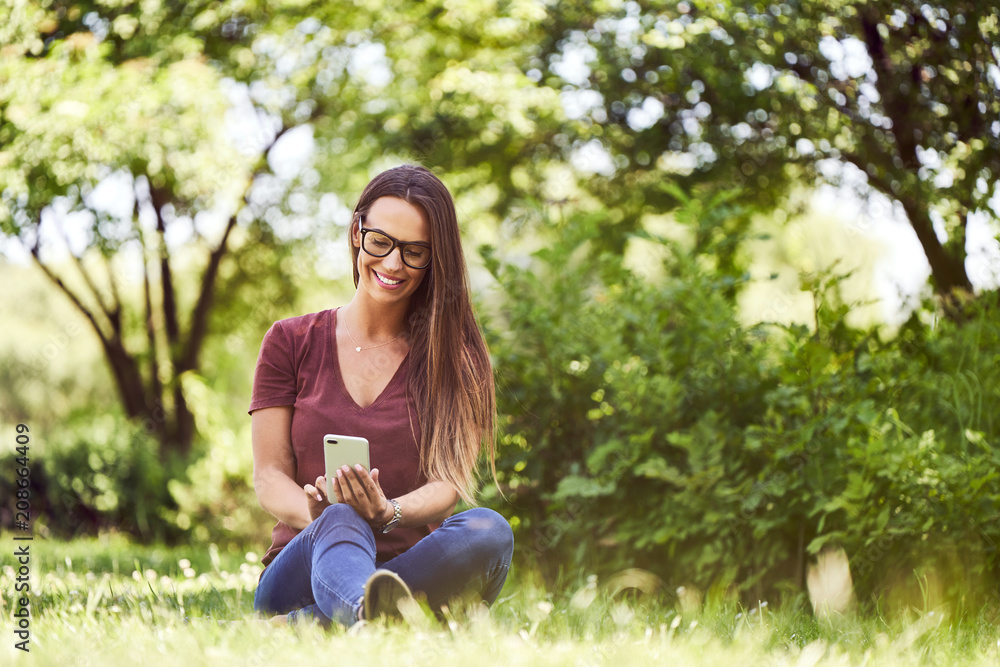 Fototapeta premium Smiling young woman sitting on ground in a park and using smartphone