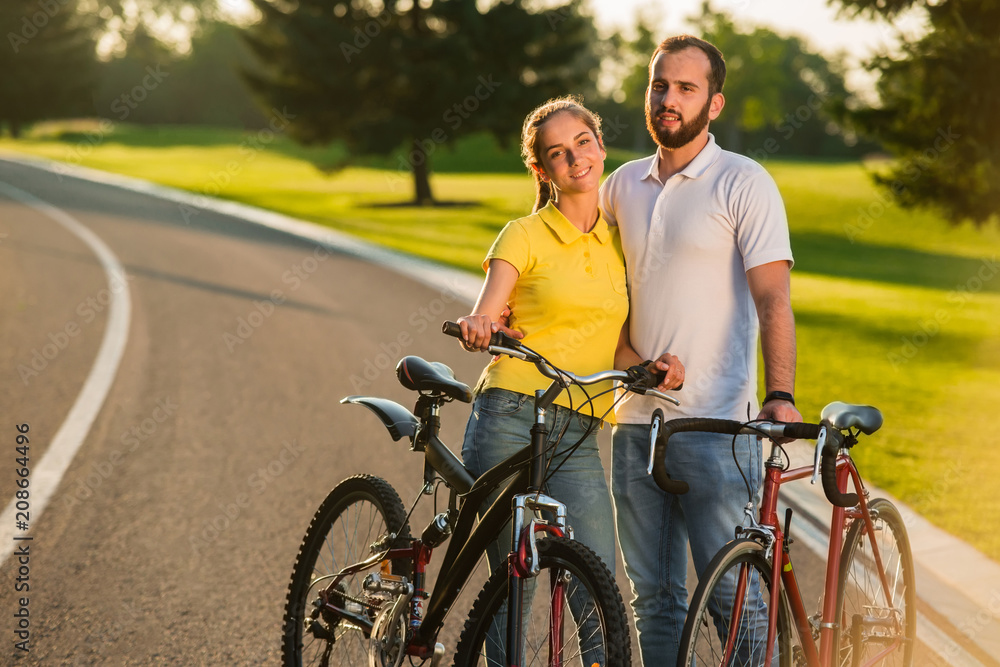 Loving couple enjoying sunny day on bikes. Young people in love standing with bicycles on the road and looking at camera. Romantic holidays concept.