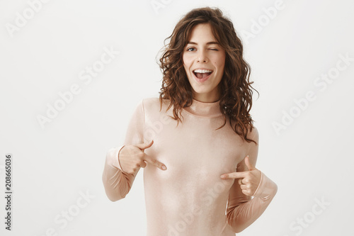 Free nipple. Portrait of carefree and confident joyful female with curly hair, covering nipples with fingers, smiling broadly and winking with flirty expression, being in sensual and romantic mood