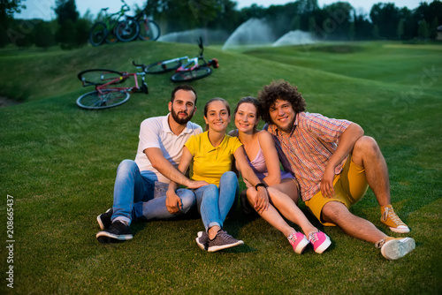Outdoors shot of cyclists resting on green grass. Group of young smiling friends sitting on the meadow and looking at camera. Evening cycling with friends.