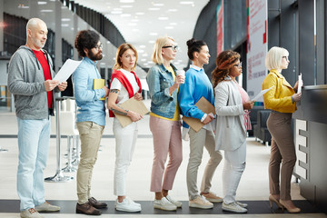 Group of modern multiethnic people of different ages wearing casual clothing holding papers and sketchpads while standing in long line for registration