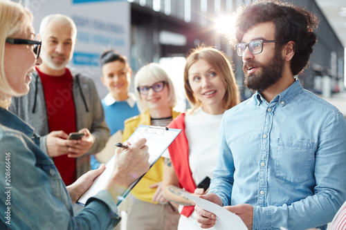 Confident hipster mixed race man with beard saying his name while holding registration for participating in forum, manager finding his name in list while other people waiting for turn