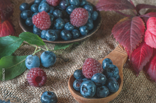 Ripe blueberries and raspberries lie in a handmade ceramic pot and a wooden spoon with a beautiful autumn leaf.