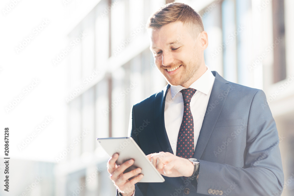 Handsome prosperous businessman reads useful information in internet via touchpad, connected to wireless internet, poses outdoor. Confident office worker makes booking online. Technology concept Handsome prosperous businessman reads useful information in internet via touchpad, connected to wireless internet, poses outdoor. Confident office worker makes booking online. Technology concept