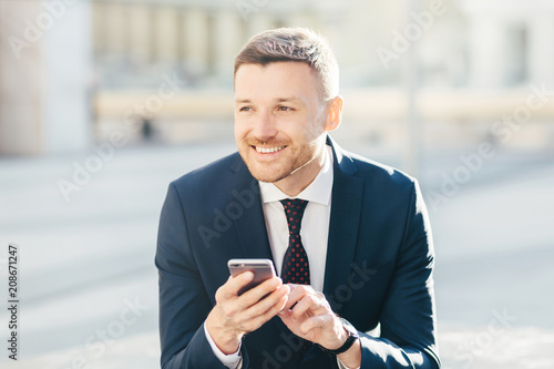 Horizontal shot of attractive male with cheerful thoughtful expression, uses modern mobile phone for searching information on internat website, connected to high speed 4G internet, makes money online photo