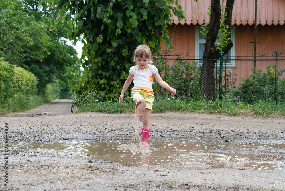 Naklejka premium Little girl is playing in the water in the middle of a ruined road, after rain