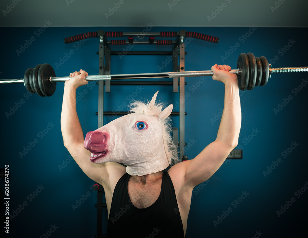 Funny man in comical mask doing sport exercise. Unusual guy pump up iron at home. Unicorn sportsman. - obrazy, fototapety, plakaty 