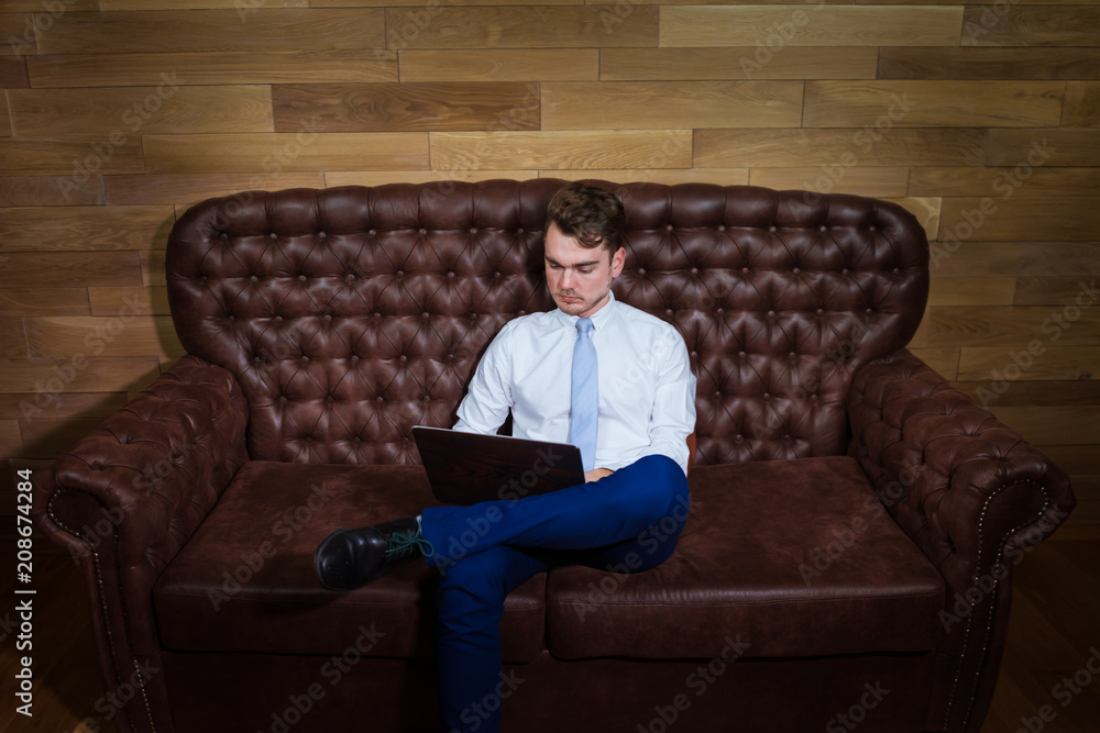 Serious man in suit is working with laptop at home office. Confident young manager sits on the leather sofa on background of wooden wall.