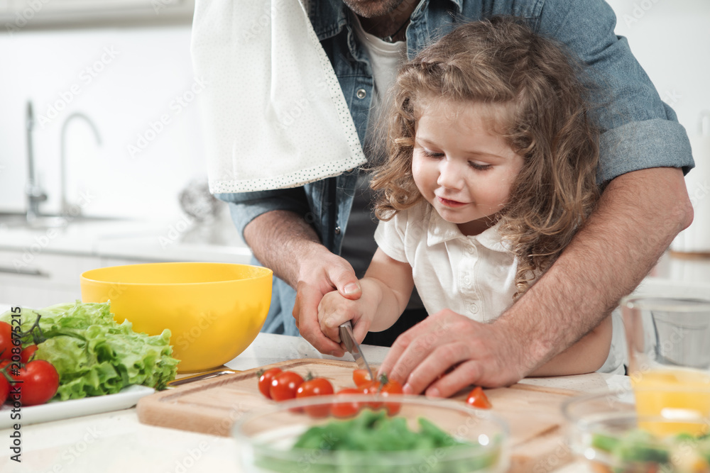 Portrait of curious daughter cutting tomato together with her father. She is laughing while standing in kitchen Portrait of curious daughter cutting tomato together with her father. She is laughing while standing in kitchen