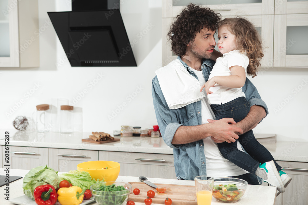 Cheerful dad is looking after his child at home. He is holding a girl in arms while cooking healthy breakfast in kitchen 