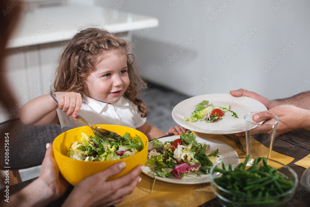 Eat, my baby. Portrait of excited little girl is eating salad while sitting at table near parents. Father and mother holding plates with healthy food in front of their daughter Eat, my baby. Portrait of excited little girl is eating salad while sitting at table near parents. Father and mother holding plates with healthy food in front of their daughter