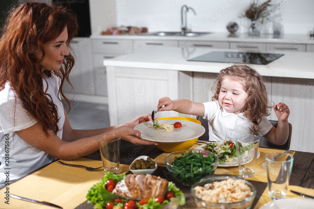 Joyful little girl is taking fresh vegetables from plate by spoon. Her mom is looking at kid with love and smiling while sitting next to her. Parenthood concept 