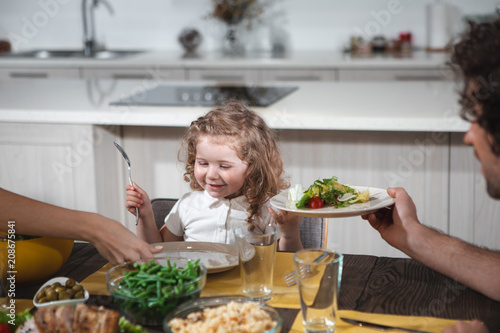 Mom and dad are feeding their little daughter by healthy food. Child is looking at plate with excitement and smiling. Family lunch concept 