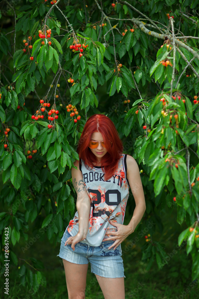 Obraz premium Beautiful woman with red hair standing in front of a cherry tree with ripe fruits. warm summer mood.