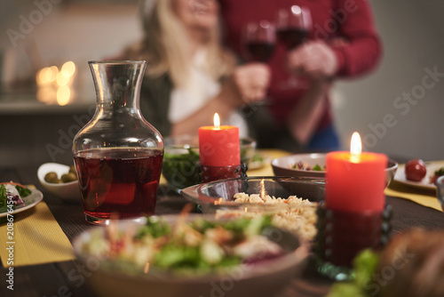 Focus of candles lit on festive dinner. There are red wine and variable dishes on table creating romantic atmosphere for couple on background 