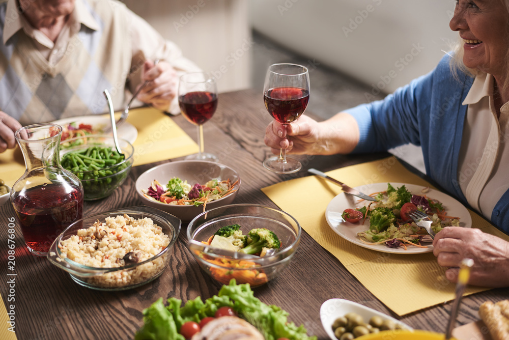 Close up of elderly woman hand holding red wine glass. She is sitting at table full of healthy dishes next to husband. Old female is raising goblet while eating salad with smile Close up of elderly woman hand holding red wine glass. She is sitting at table full of healthy dishes next to husband. Old female is raising goblet while eating salad with smile