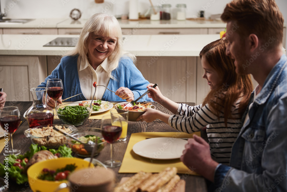 Three generations at one table. Happy grandmother talking to her dearest. Small girl is holding bowl with salad while her dad is sitting next to her. They are eating wholesome meal with joy Three generations at one table. Happy grandmother talking to her dearest. Small girl is holding bowl with salad while her dad is sitting next to her. They are eating wholesome meal with joy