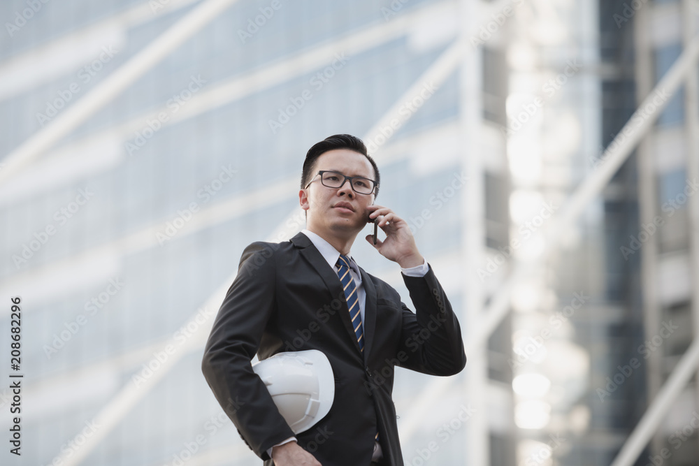 Portrait of a young asian businessman talking on mobile phone and holding a helmet. Man outdoor with phone in a city Bangkok Thailand. Concept of communication Portrait of a young asian businessman talking on mobile phone and holding a helmet. Man outdoor with phone in a city Bangkok Thailand. Concept of communication
