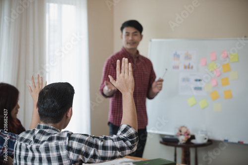 A young Asian man wearing a plaid shirt, lifting a hand to ask questions and, in a conference room, a business.