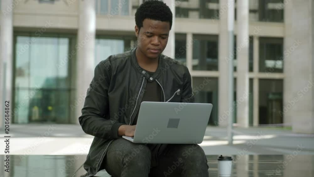 Medium shot of young African American man in leather jacket typing on laptop outdoors, looking away thoughtfully and nodding