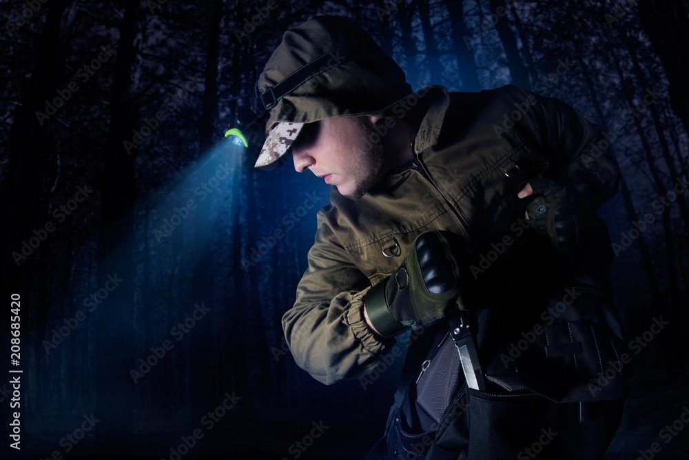 Profile view photo of a male person in brown tactical outfit jacket, gloves, backpack and head flashlight taking out knife on night woods background. - obrazy, fototapety, plakaty 