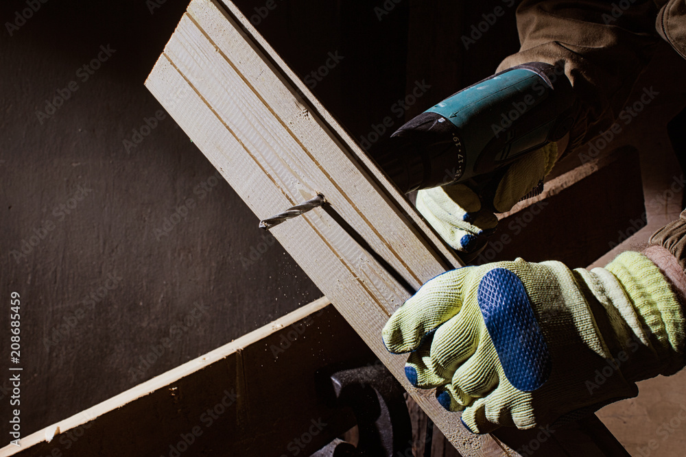 Photo of a carpenter worker in yellow and blue gloves and outfit drilling a hole in wooden board with electric drill on black background. Photo of a carpenter worker in yellow and blue gloves and outfit drilling a hole in wooden board with electric drill on black background.