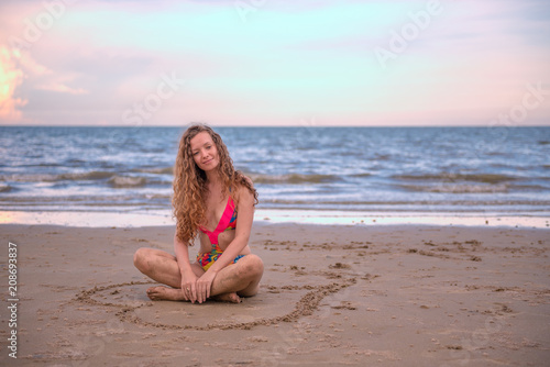 Young healthy woman with blonde curly hair sitting in a hand writing of heart shaped on the sand, summer beach by the sea with waves, blue sky, relaxing and happy time concept.