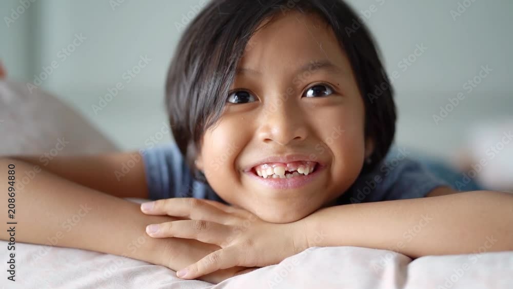 Cute little girl smiling at the camera with her first lost milk tooth, lying on the bedroom while watching TV at home Cute little girl smiling at the camera with her first lost milk tooth, lying on the bedroom while watching TV at home