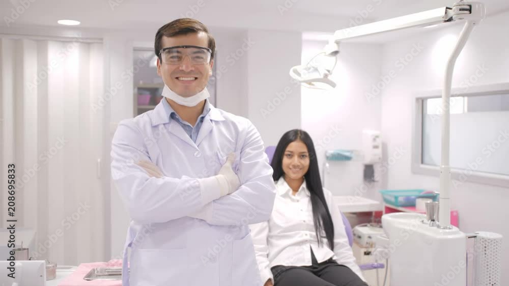 Portrait of smiling caucasian dentist in dental studio with young asian woman patient smiling in background. Portrait of smiling caucasian dentist in dental studio with young asian woman patient smiling in background.