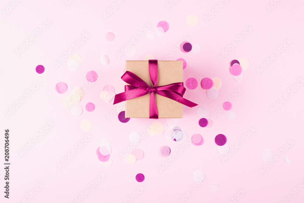 Kraft gift box tied with pink bow on pink background decorated with confetti.. Top view, holiday present concept.