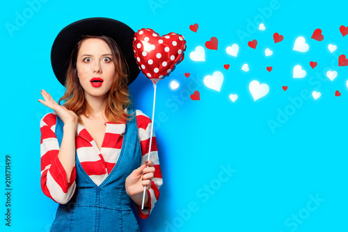 suprised red-haired white european woman in hat and red striped shirt with jeans dress with heart shape toy on blue background with hearts