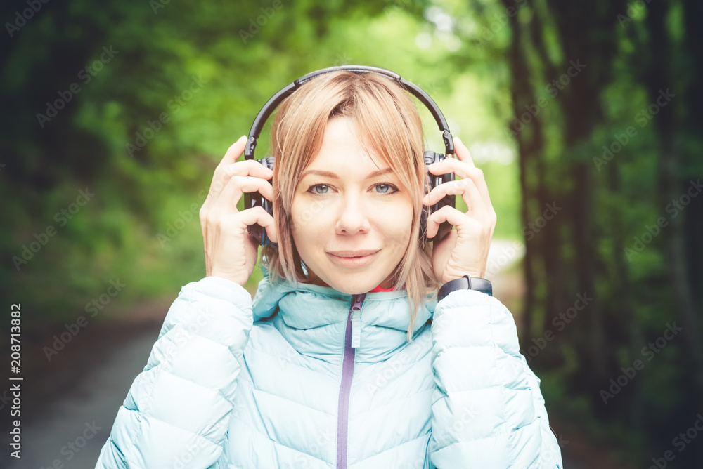 Portrait of an attractive sports girl blonde in a light running down jacket dressing bluetooth headphones with music or the sounds of nature while on a forest road Portrait of an attractive sports girl blonde in a light running down jacket dressing bluetooth headphones with music or the sounds of nature while on a forest road