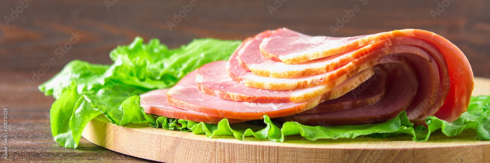 Sliced ham with fresh green lettuce leaves on a round cutting board. Meat products on a brown wooden table. banner Sliced ham with fresh green lettuce leaves on a round cutting board. Meat products on a brown wooden table. banner