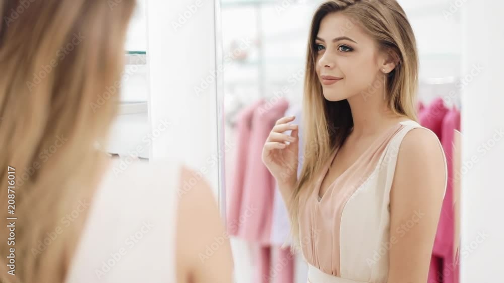 Beautiful shopping woman trying dress near mirror in room. Young attractive girl in a dressing room at a shopping center Clothing, fashion, wardrobe, style and people concept Beautiful shopping woman trying dress near mirror in room. Young attractive girl in a dressing room at a shopping center Clothing, fashion, wardrobe, style and people concept