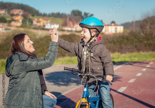 Happy mother and son giving five by the success in the learn to ride a bicycles in the city on a sunny winter day. Family leisure outdoors concept.