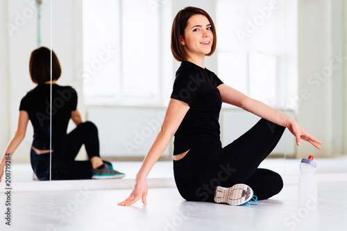 Pretty athletic girl with short brown hair in black sportswear sitting on the floor in white interior yoga or pilates studio. Mirror reflection behind her.