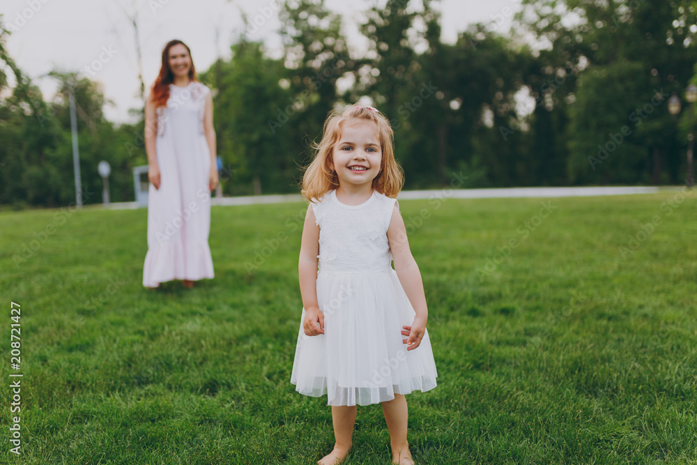 Smiling little cute child baby girl in light dress walking, play and have fun on green grass lawn in park. Mother, little kid daughter. Mother's Day, love family, parenthood, childhood concept. Smiling little cute child baby girl in light dress walking, play and have fun on green grass lawn in park. Mother, little kid daughter. Mother's Day, love family, parenthood, childhood concept.