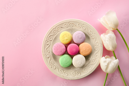 Plate with tasty macarons and flowers on color background, top view