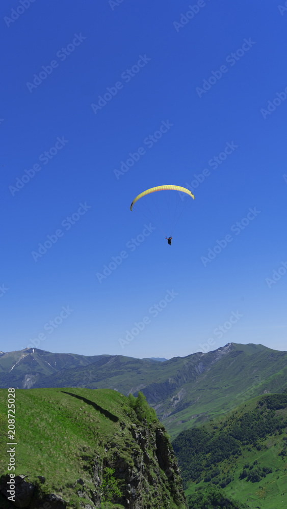 A paraglider with people flies over the mountain peaks in a bright blue sky.