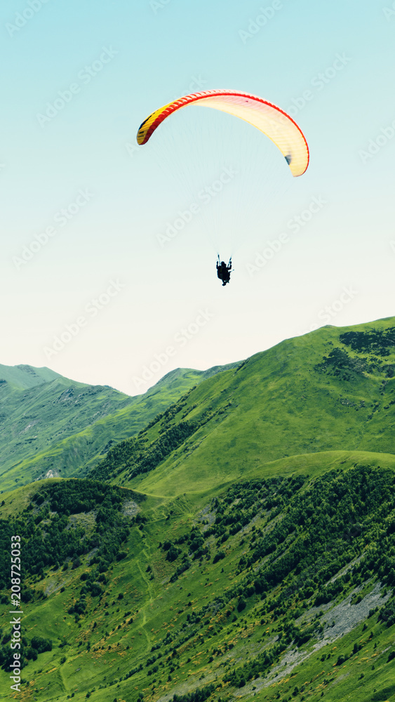 A paraglider with people flies over the mountain peaks in a bright blue sky. A paraglider with people flies over the mountain peaks in a bright blue sky.