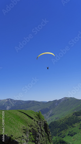 A paraglider with people flies over the mountain peaks in a bright blue sky.