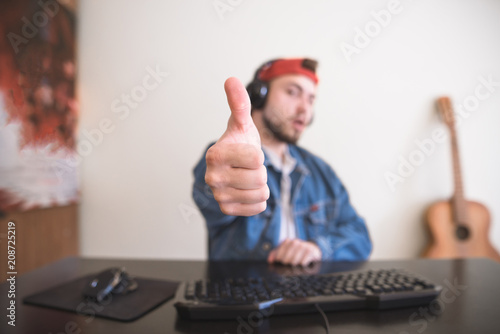 Gamer in the cap and headphones sits at home on the computer and shows the thumb up. Focus on your finger. Gamer concept photo