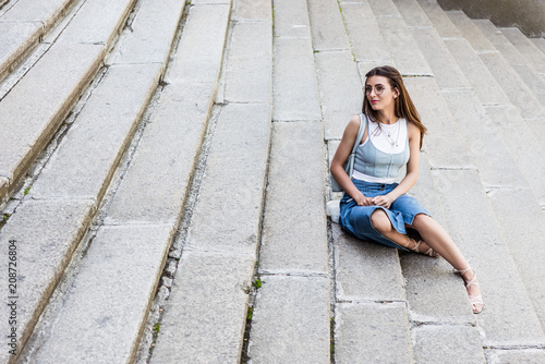 young stylish woman in eyeglasses and denim skirt resting on steps on street
