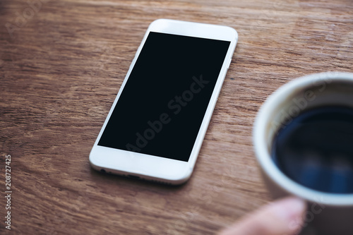 Mockup image of a hand holding coffee cup with white mobile phone with blank desktop screen on wooden table