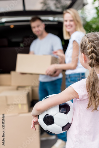 cropped shot of little child holding soccer ball while her parents unpacking cardboard boxes for relocation