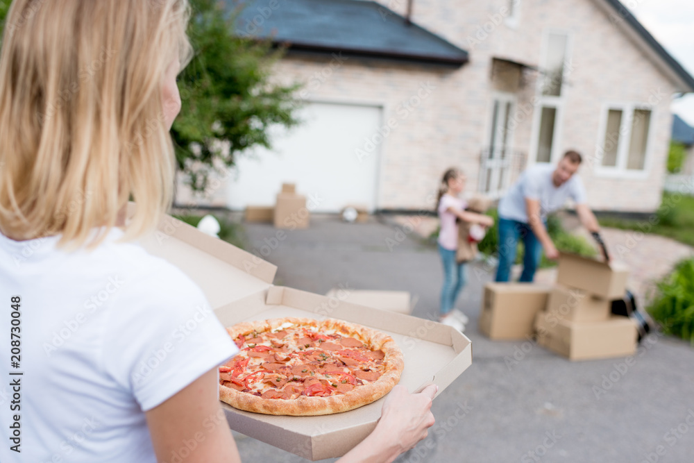 partial view of  woman holding pizza while her husband and daughter unpacking cardboard boxes in front of new house