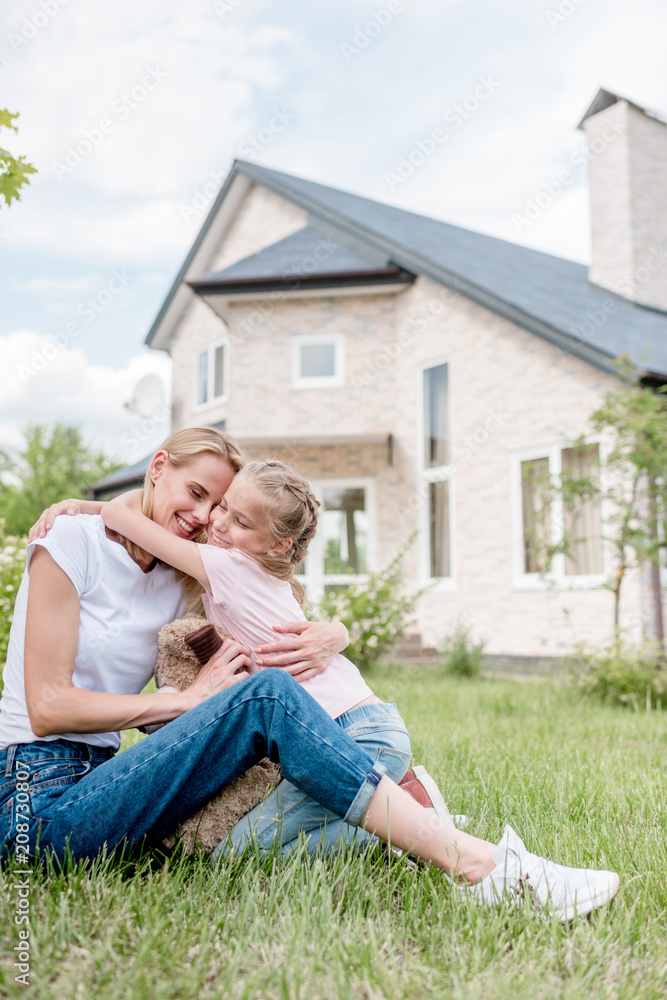 Φωτογραφία little daughter hugging smiling mother on green lawn in front of cottage