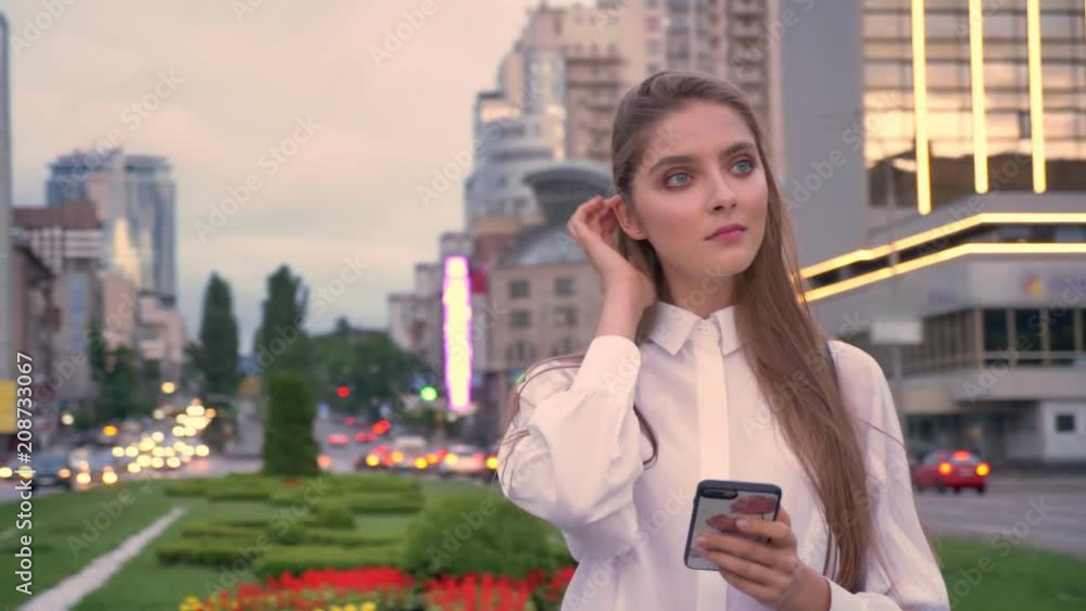 Young beautiful serious girl is typing message on her smartphone at sunset in city center in summer, thinking process, communication concept Young beautiful serious girl is typing message on her smartphone at sunset in city center in summer, thinking process, communication concept