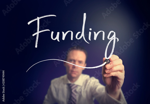 A businessman writing a Funding concept with a white pen on a clear screen.