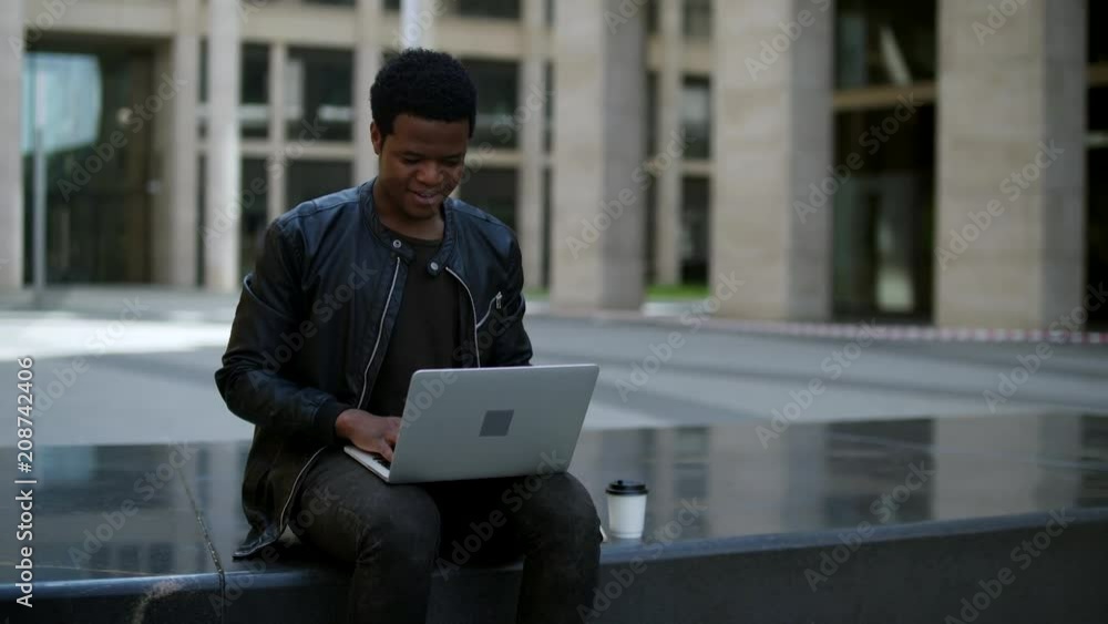 Young African American journalist or blogger sitting outdoors with paper coffee cup near him, creating new article and typing it on laptop Young African American journalist or blogger sitting outdoors with paper coffee cup near him, creating new article and typing it on laptop